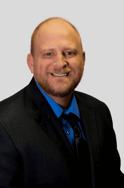 A man with a reddish beard and short hair is wearing a black suit jacket over a blue shirt and black tie, smiling at the camera against a plain light background.